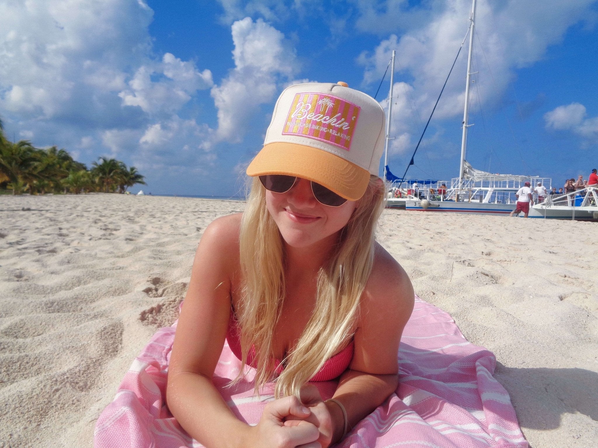 Woman wearing peach and cream vintage trucker hat with "Beachin'" text on beach with sailboats in background.