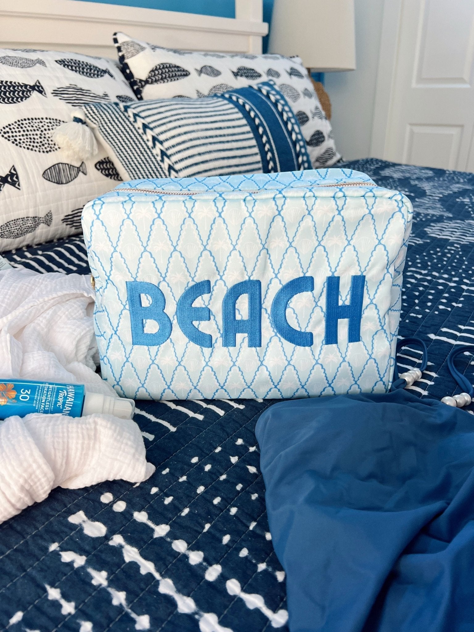 Blue and white quilted cosmetic bag with "BEACH" text, displayed on bedding with coastal patterns.