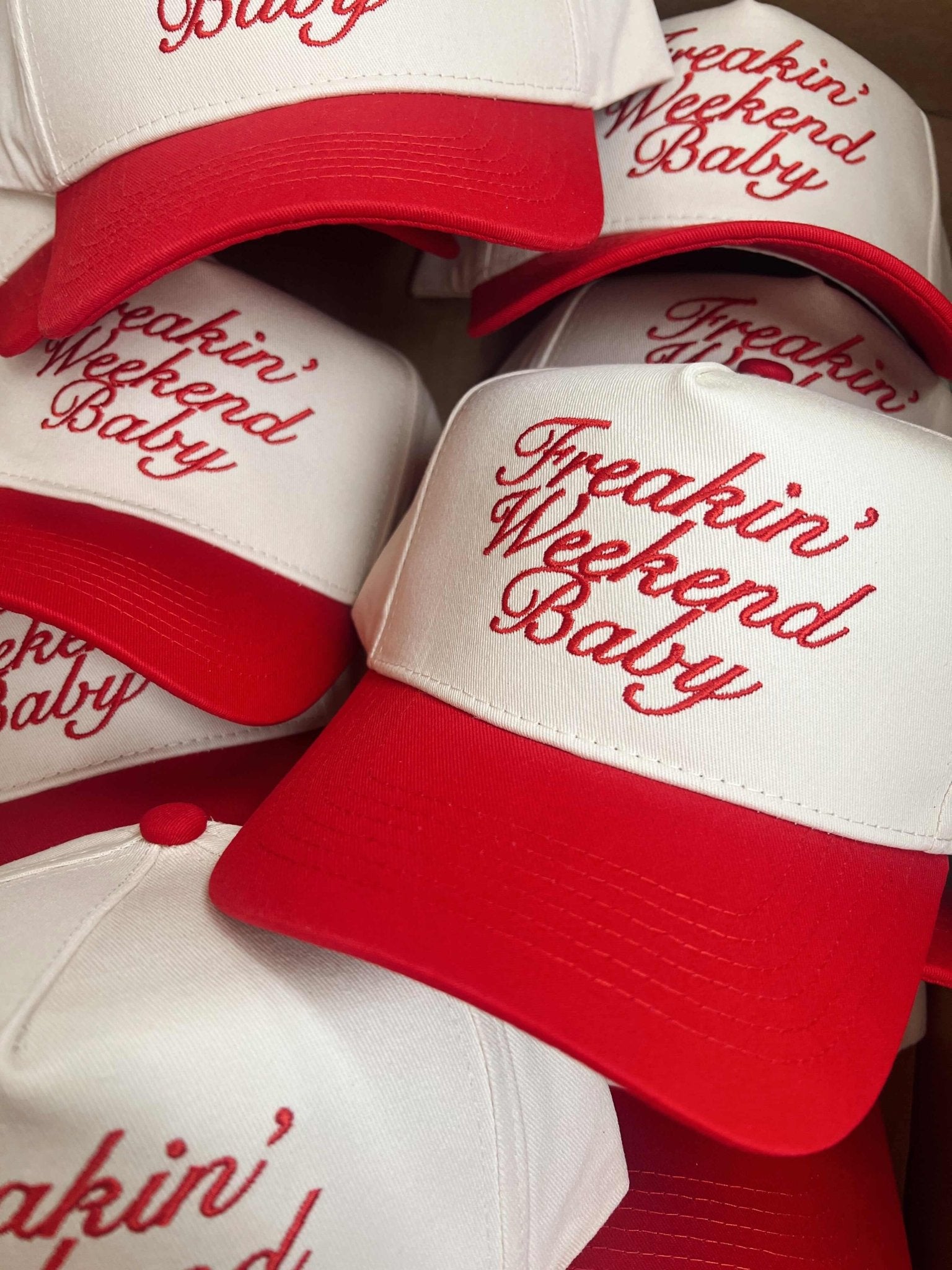 Red and white trucker hats with "Freakin' Weekend Baby" embroidered in red script lettering, stacked together showing the vintage curved bill style.
