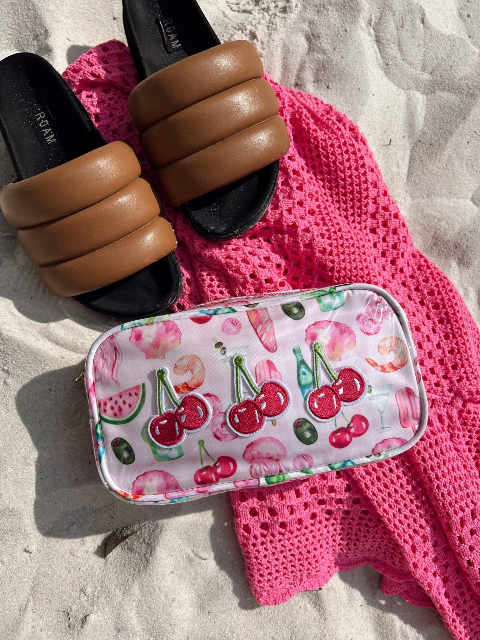 Clear cosmetic bag with cherry and watermelon print on pink knit blanket beside tan and black slides