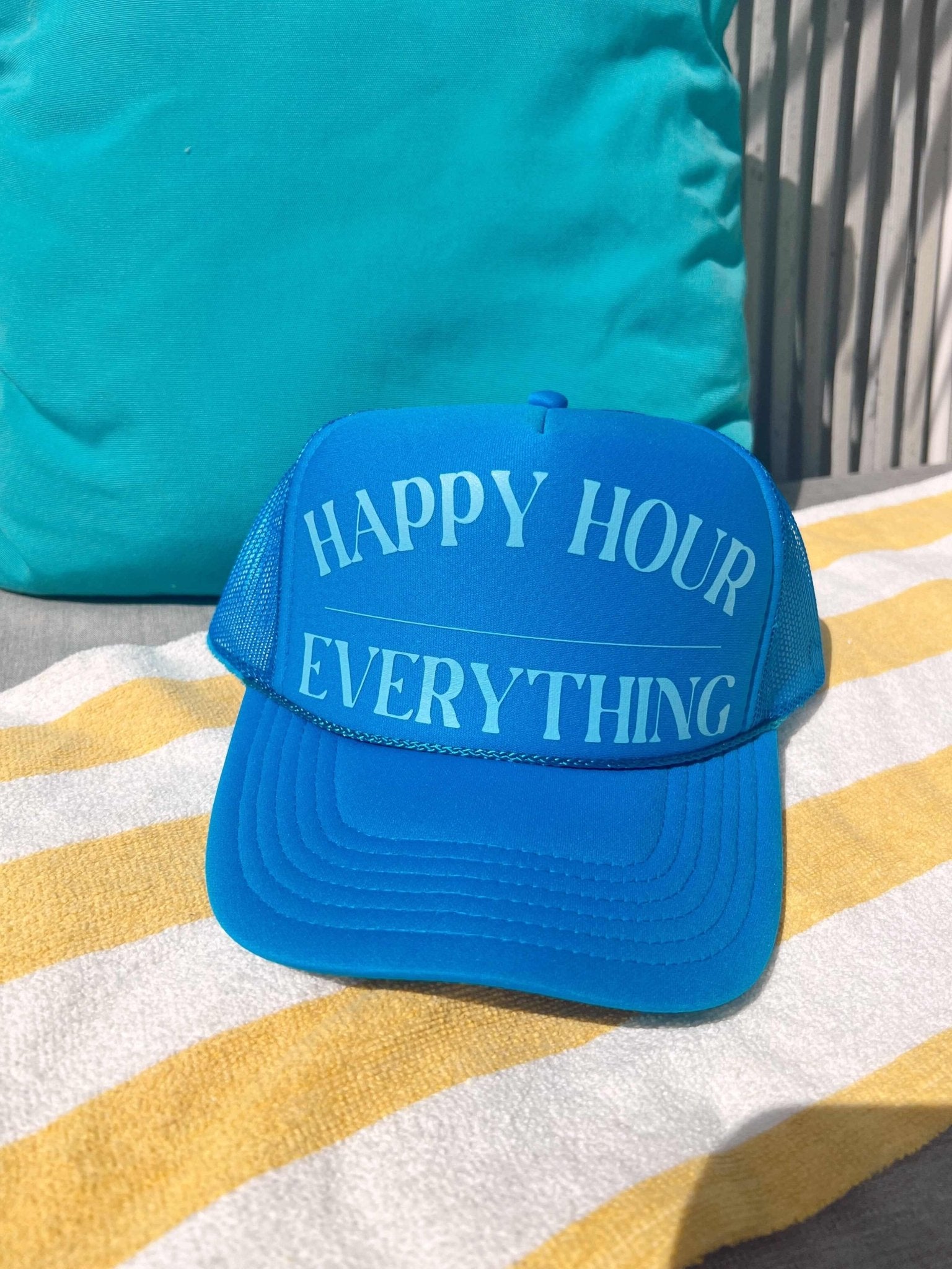 Blue trucker hat with "HAPPY HOUR EVERYTHING" text printed across the front, displayed on a striped beach towel