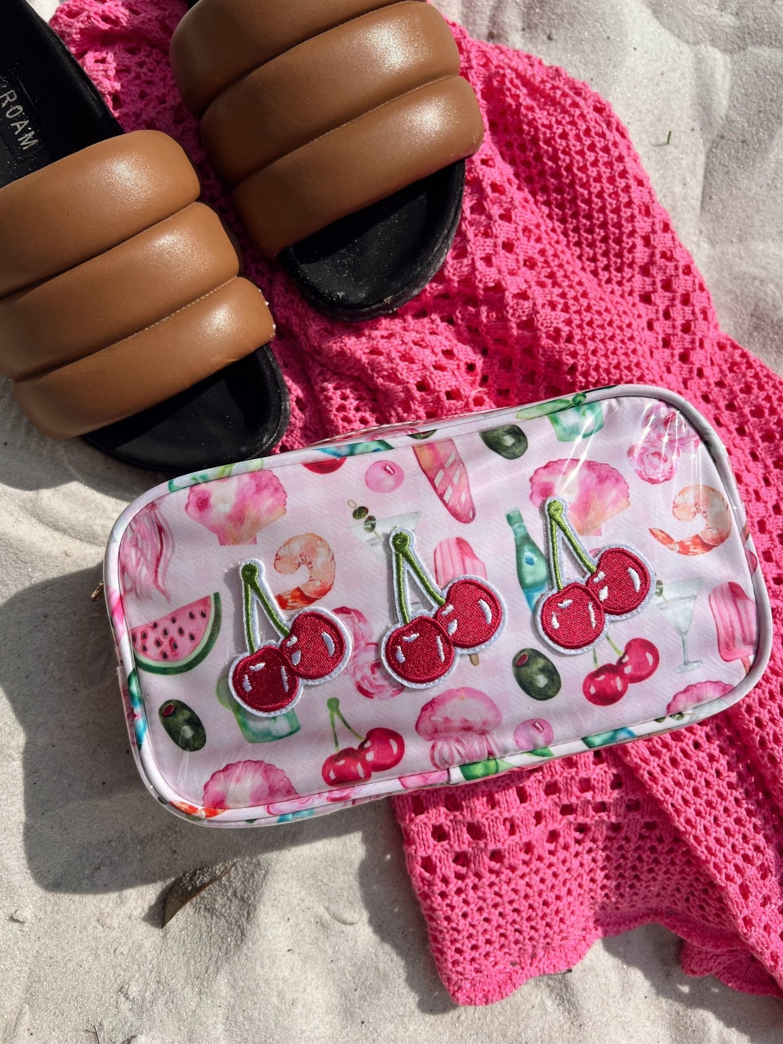 Pink cherry and watermelon print cosmetic bag on beach towel with sunscreen and sunglasses nearby.