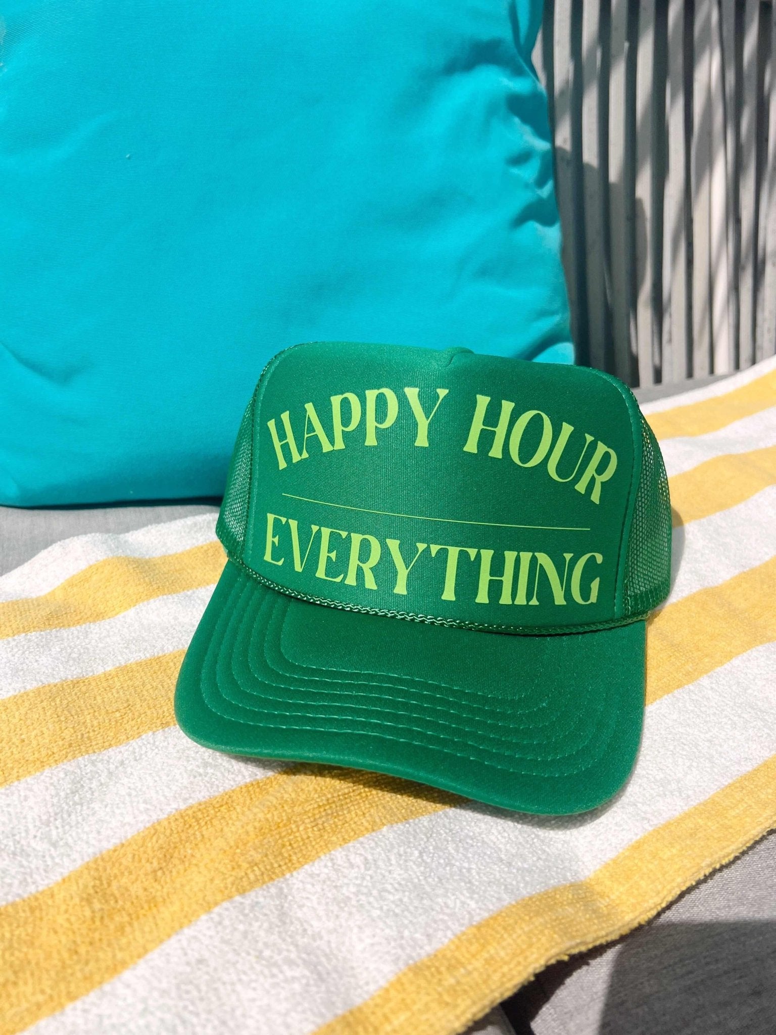 Green trucker hat with yellow embroidered text reading "Happy Hour" and "Everything" on striped beach towel background