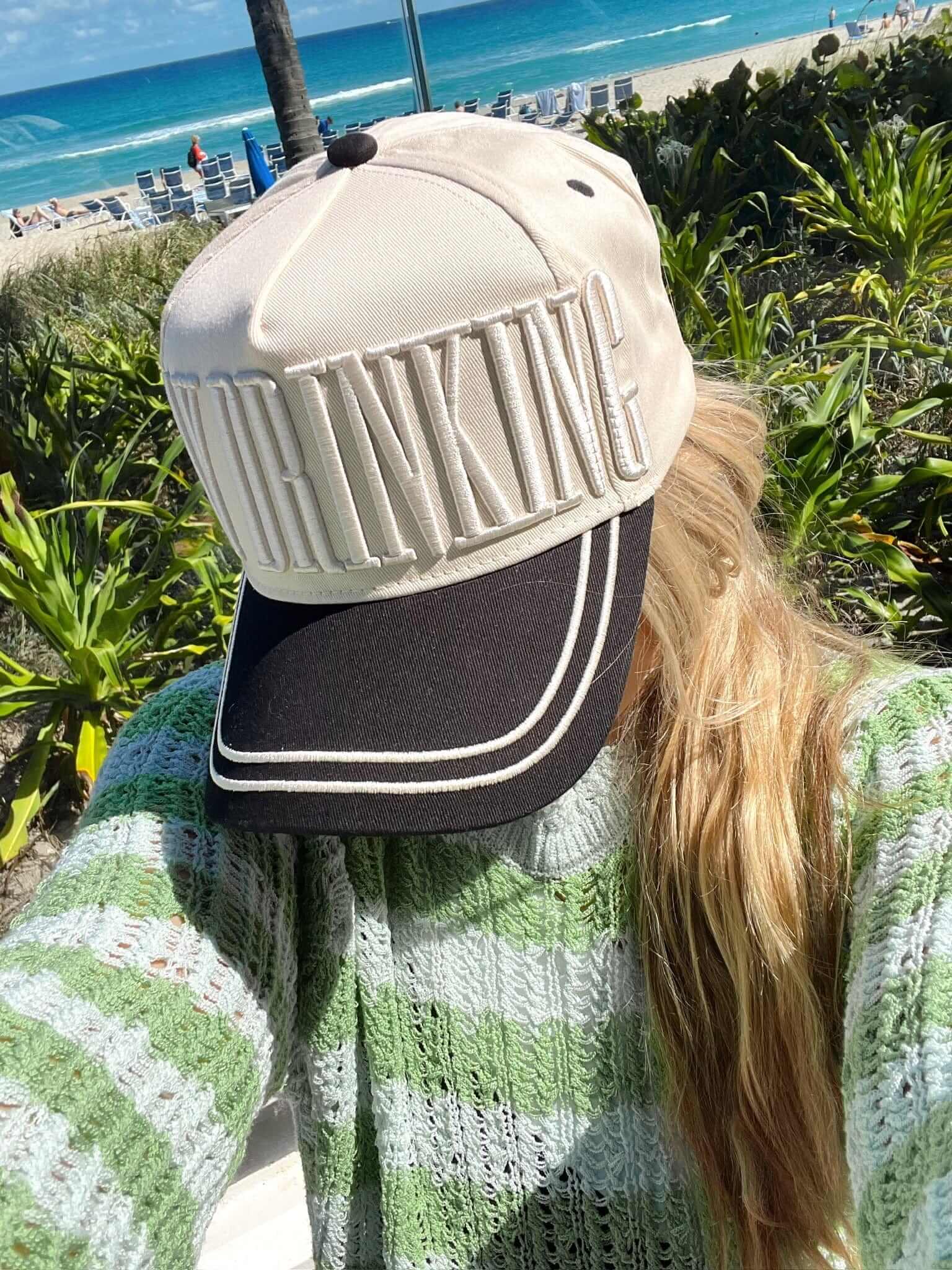 Black and cream vintage trucker hat with mesh back panel and white stripe detail, displayed on a beach setting backdrop.