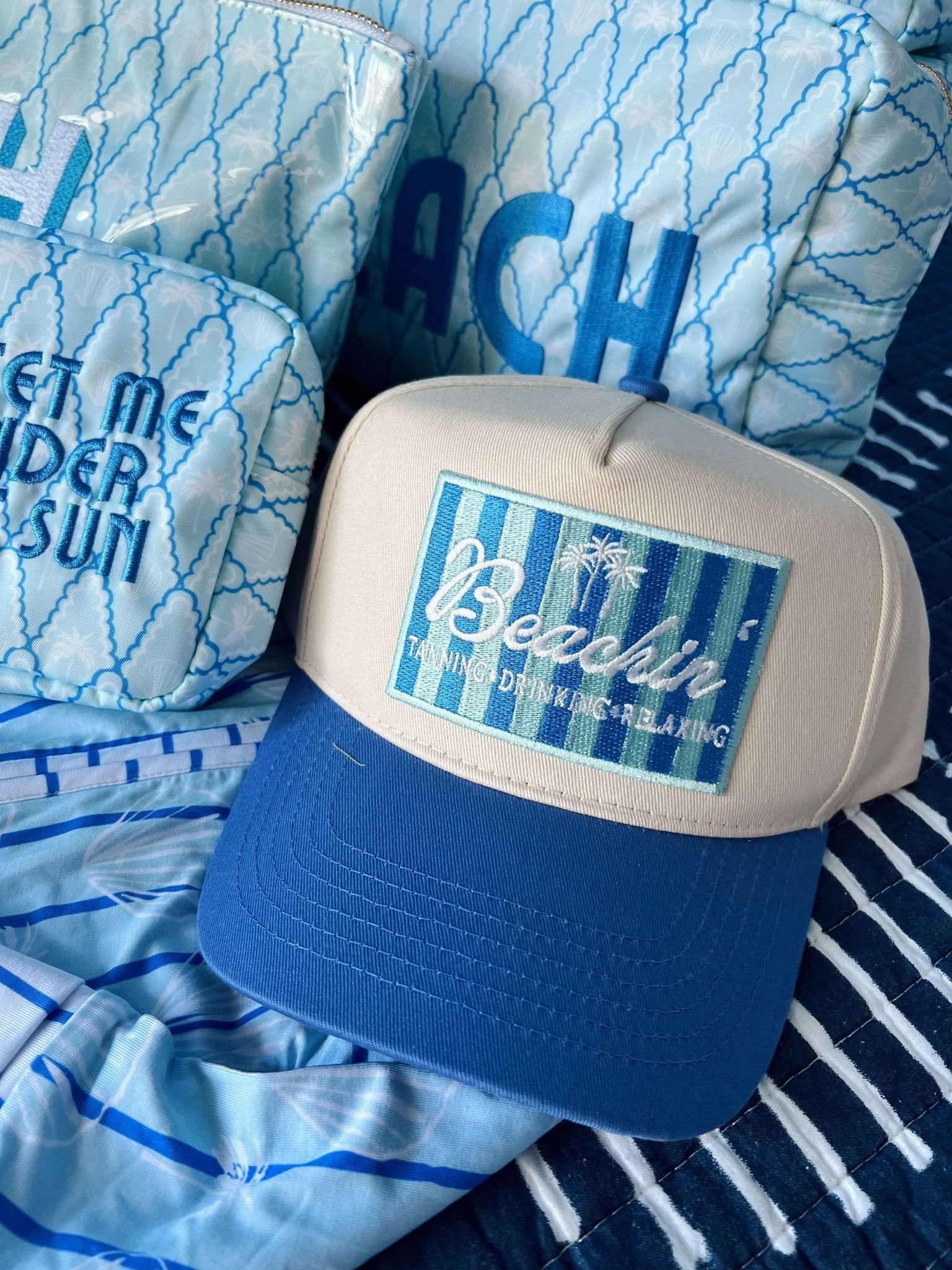 Blue and white vintage trucker hat with "Beachin'" text and palm tree design on front panel, displayed on beach-themed fabric.