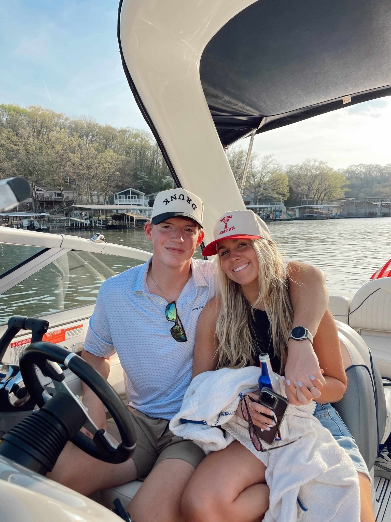 Couple sitting on a boat wearing white trucker hats with "DRUNK" text and vintage styling, water and trees in background.