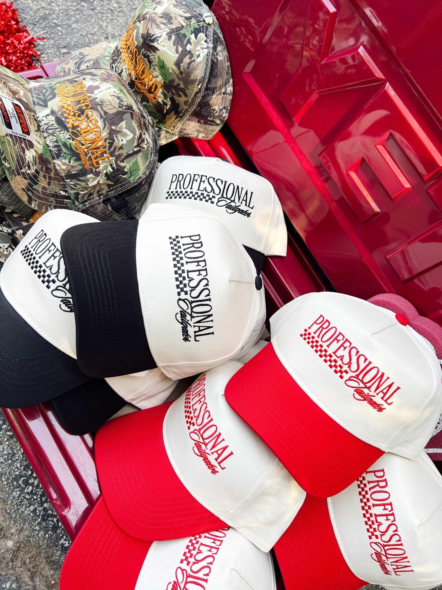 Assorted trucker hats with "Professional Tailgater" text in red and black colorways, displayed in a pink cooler with metallic balloon decorations.
