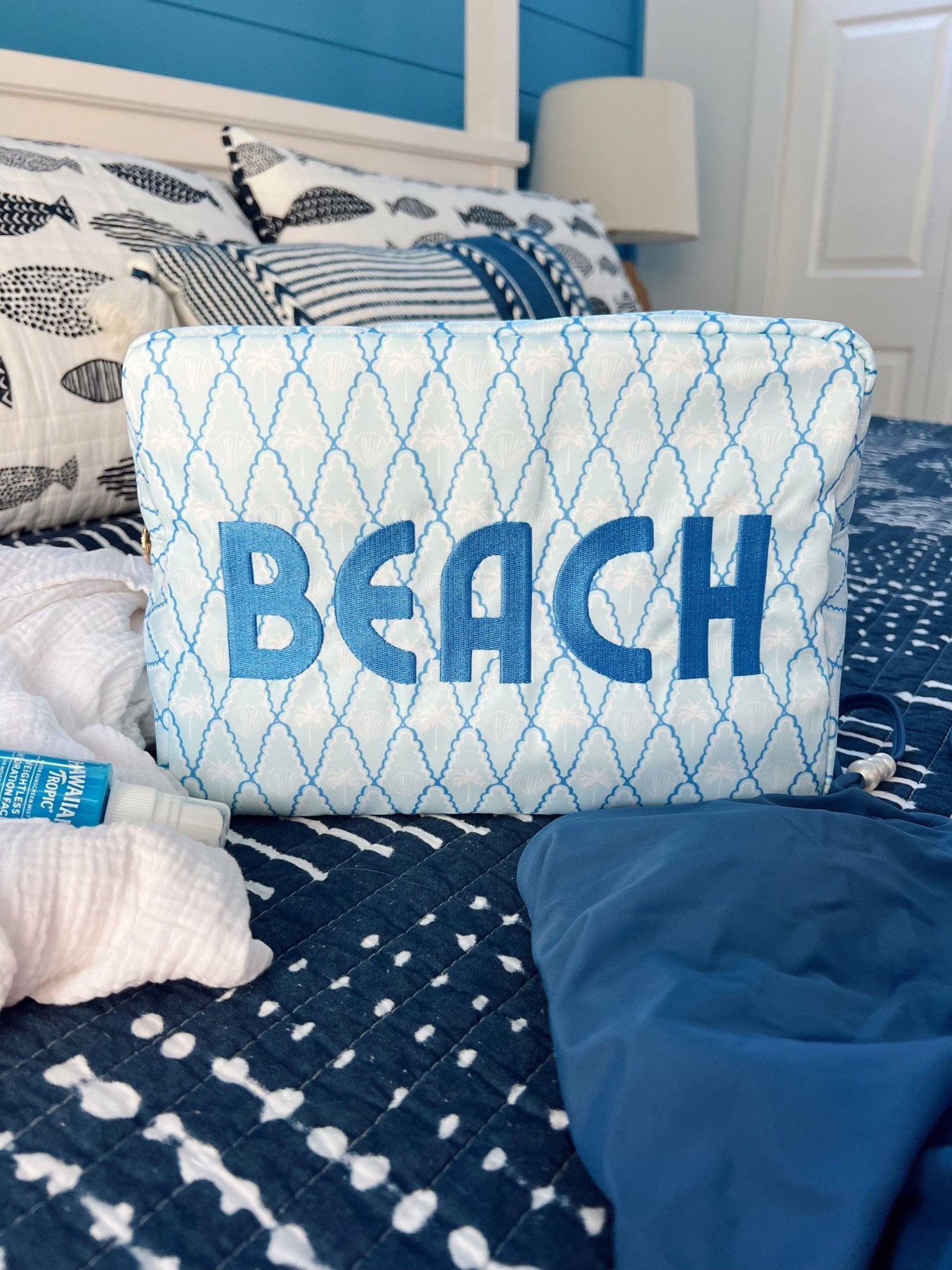 Blue and white geometric patterned makeup bag with "BEACH" text, displayed on bedding with coastal decor.
