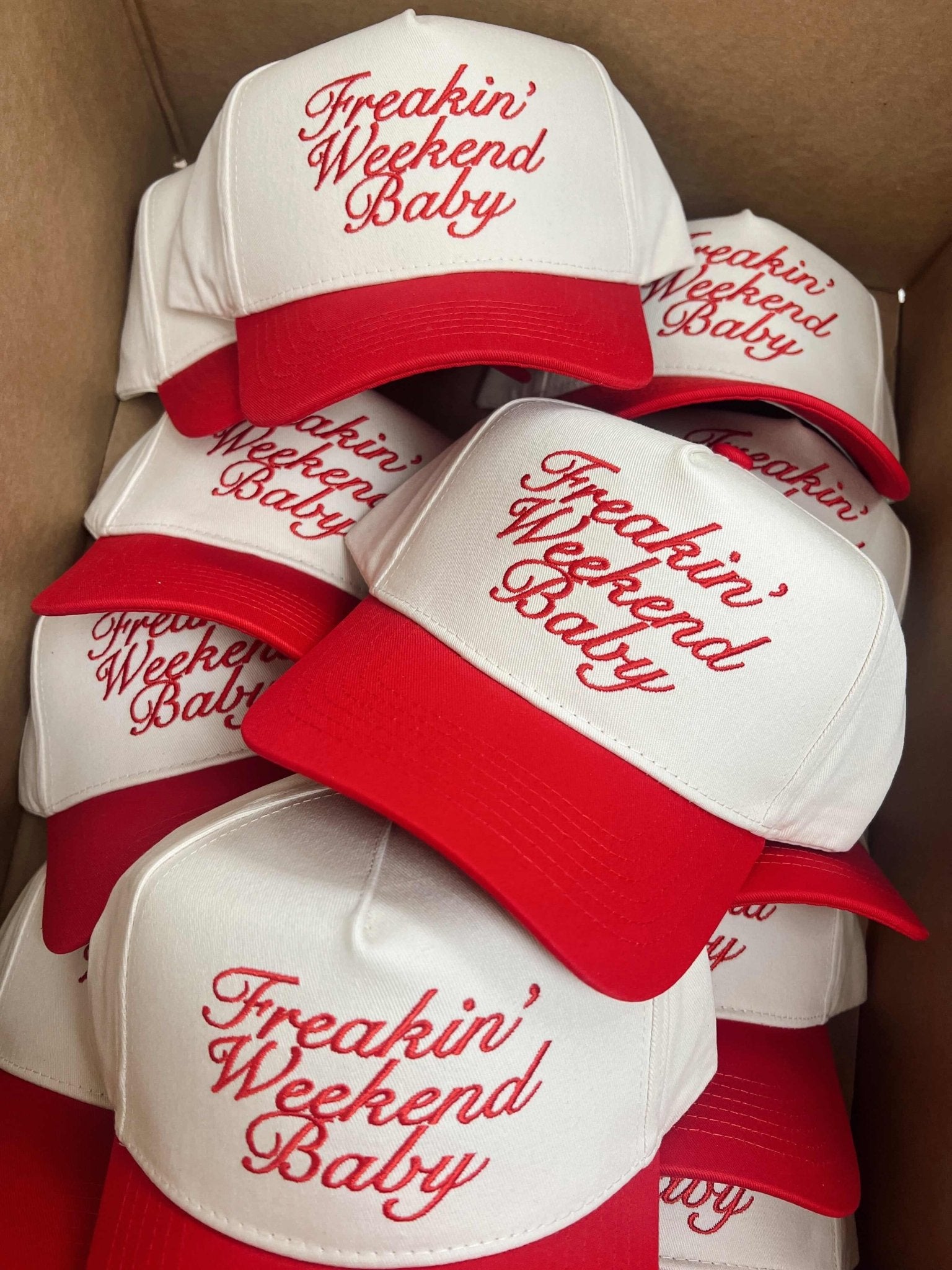 Stack of white trucker hats with red visor and "Freakin' Weekend Baby" text in red script lettering