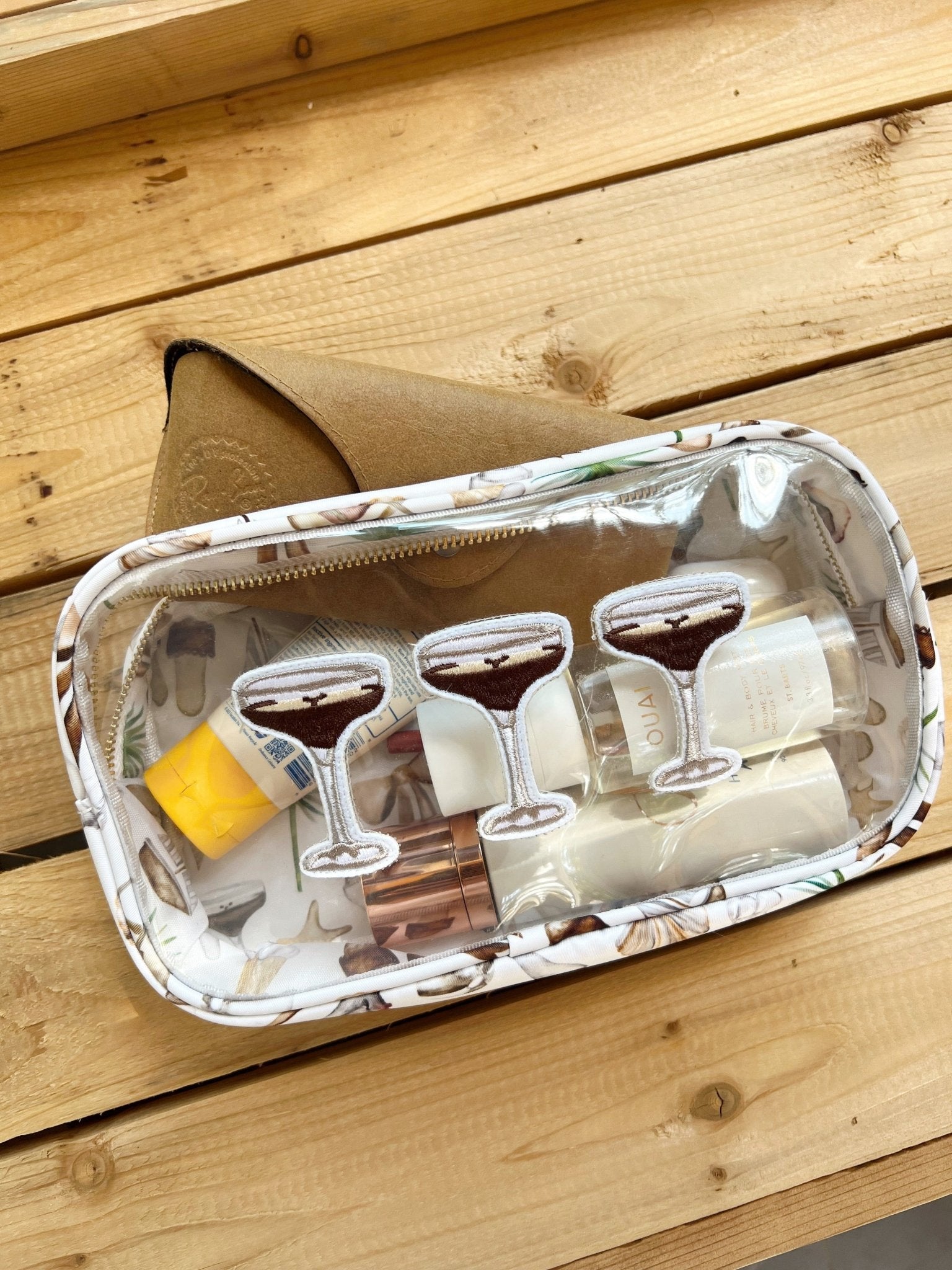 Clear travel bag with neutral coastal pattern containing two martini glasses, cocktail accessories, and yellow sponge on wooden surface.