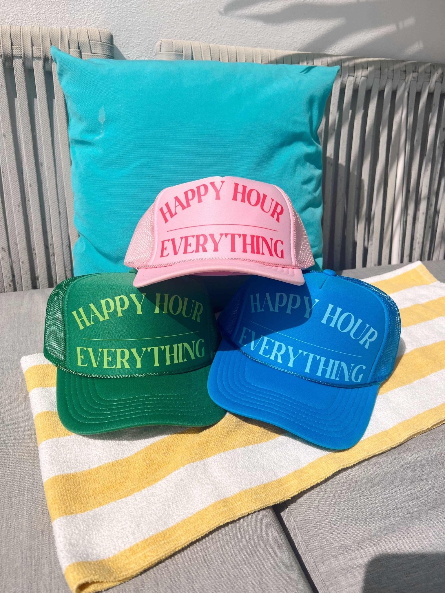 Turquoise, green, and blue trucker hats with "Happy Hour Everything" text displayed on striped beach towel