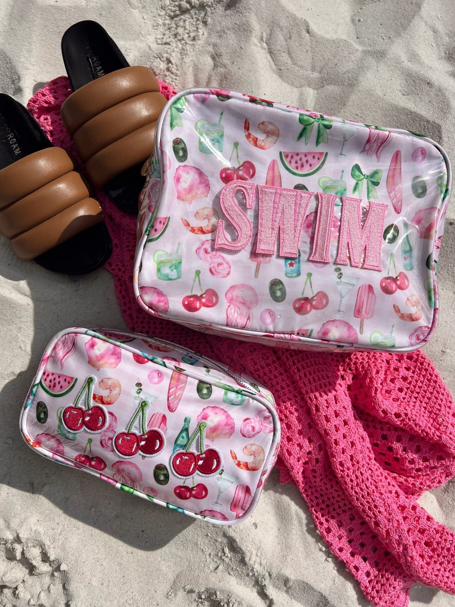 Pink fruit print travel bag with "SHINE" text, paired with matching small pouch, on sandy beach with pink accessories.