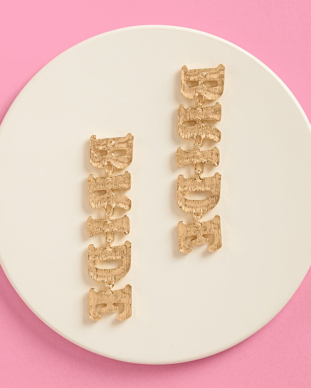 Pair of gold bride drop earrings with decorative textured design on cream round dish against pink background