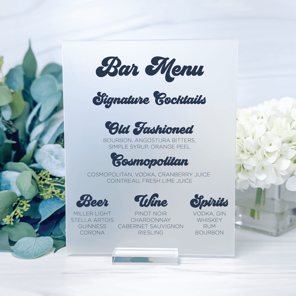 White acrylic bar menu sign with navy script text listing signature cocktails, beer, wine, and spirits, flanked by greenery and white flowers.