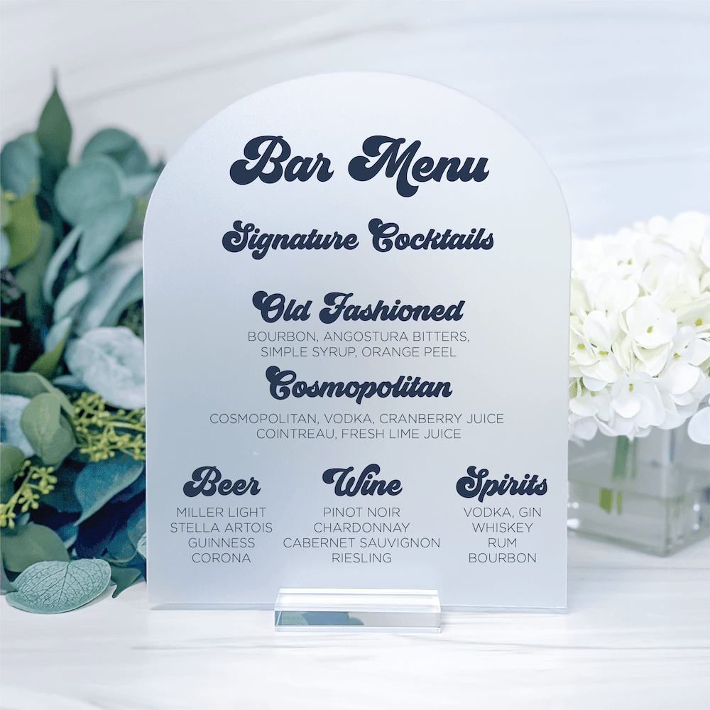 White arch-shaped bar menu sign with navy script and printed cocktail listings, displayed with greenery and white flowers.