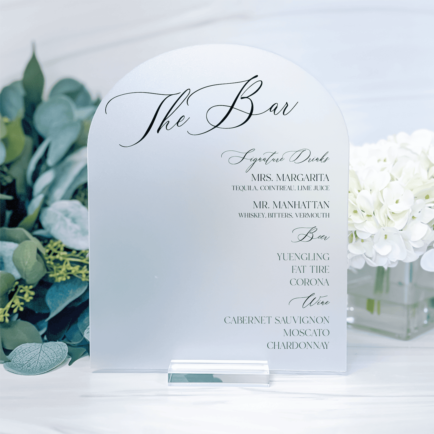 White acrylic bar menu sign with elegant script text listing drink names and cocktail options, displayed with eucalyptus and white flowers.