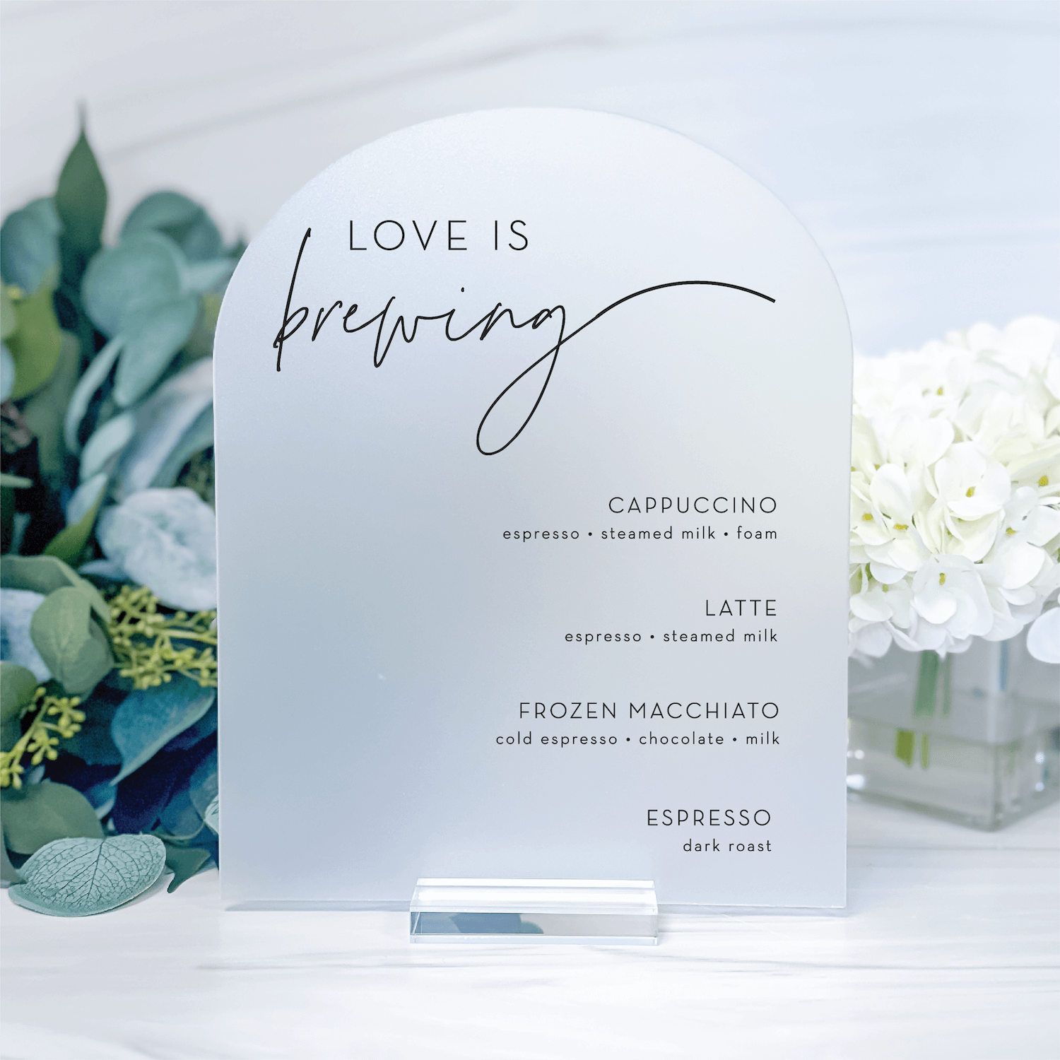 White frosted acrylic table sign with "Love Is Brewing" text and coffee drink menu options, displayed with eucalyptus and white flowers.