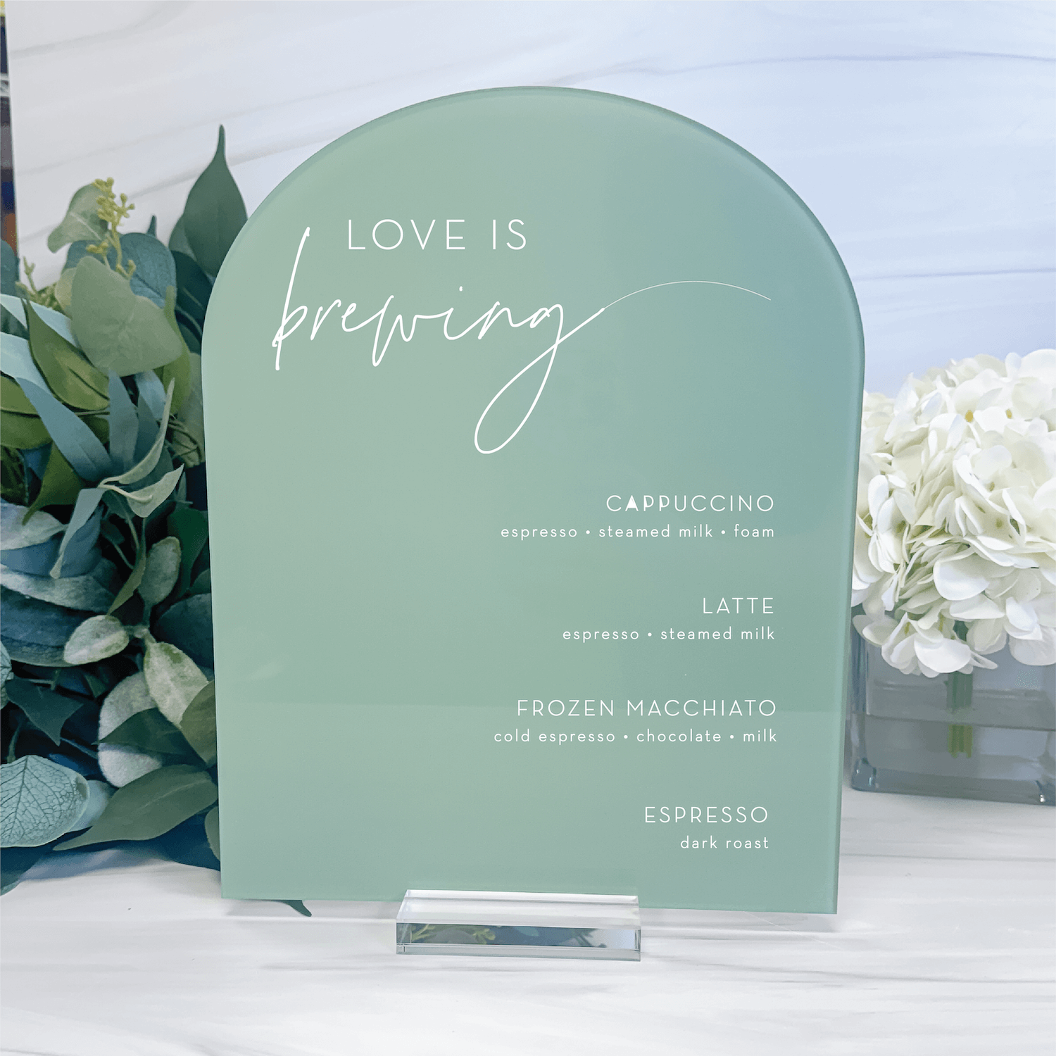 Sage green arch-shaped acrylic sign with white script text reading "Love Is Brewing" and coffee drink options listed below, displayed with greenery and white flowers