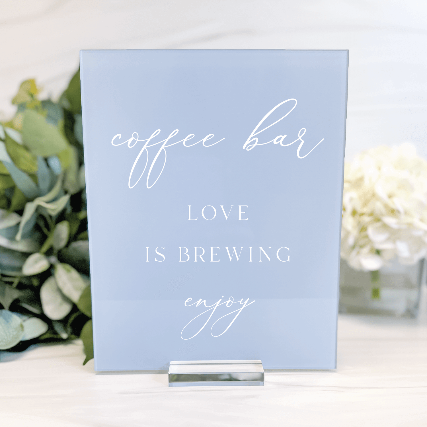 Blue acrylic sign with white script text reading "coffee bar, LOVE IS BREWING, enjoy" displayed on a stand