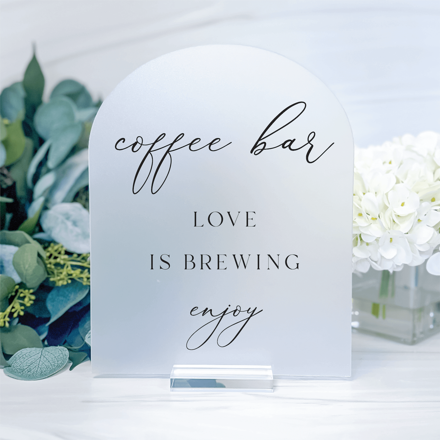 White acrylic wedding sign with "coffee bar," "LOVE IS BREWING," and "enjoy" text in script and block lettering, displayed with greenery and white flowers.