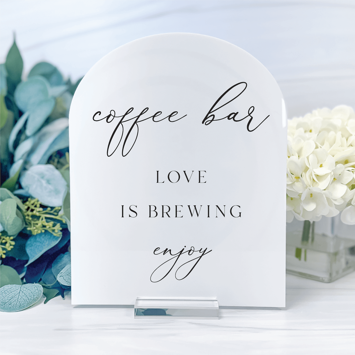 White acrylic arch sign with "coffee bar," "love is brewing," and "enjoy" text in black script and caps lettering