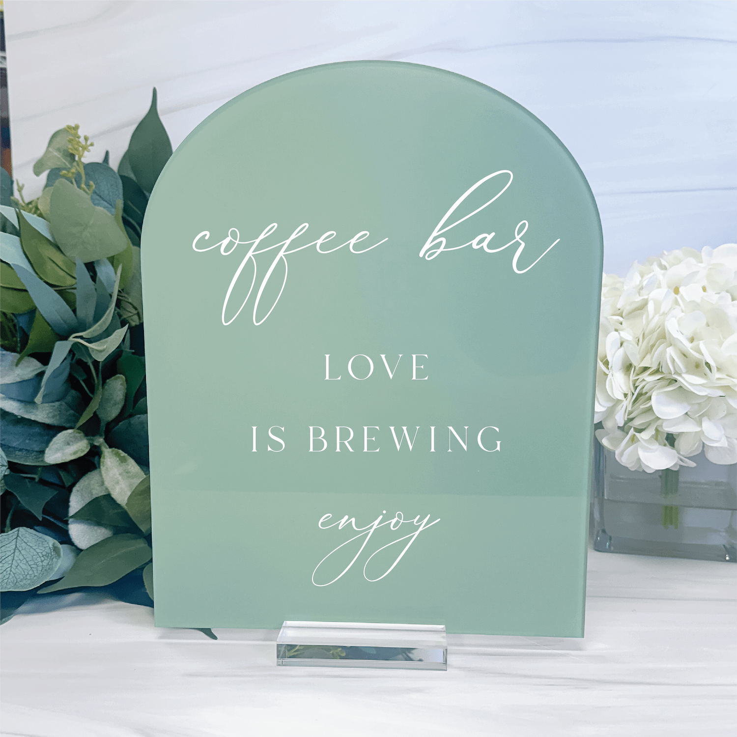 Arched sage green acrylic sign with white script and capital letters reading "coffee bar," "LOVE IS BREWING," and "enjoy," displayed with greenery and white flowers.