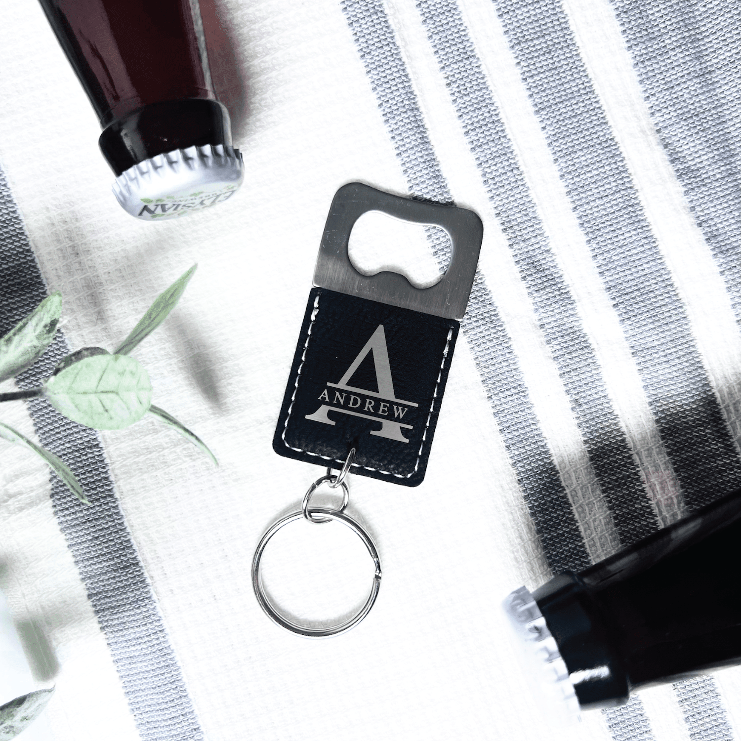 Black bottle opener keychain with personalized mountain design and metal ring, displayed on striped cloth with greenery
