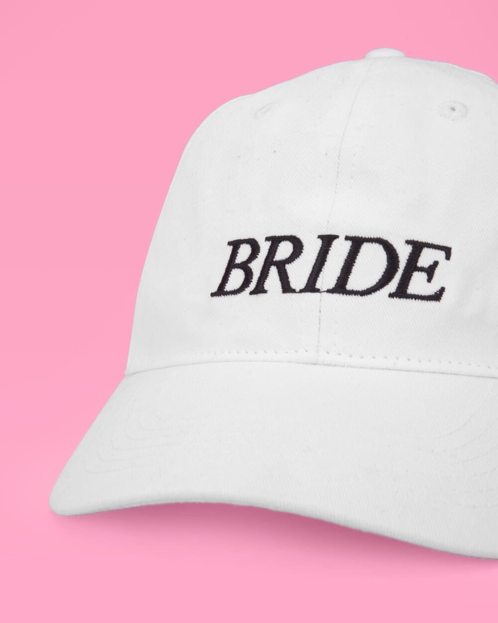 White baseball cap with black embroidered "BRIDE" text on pink background