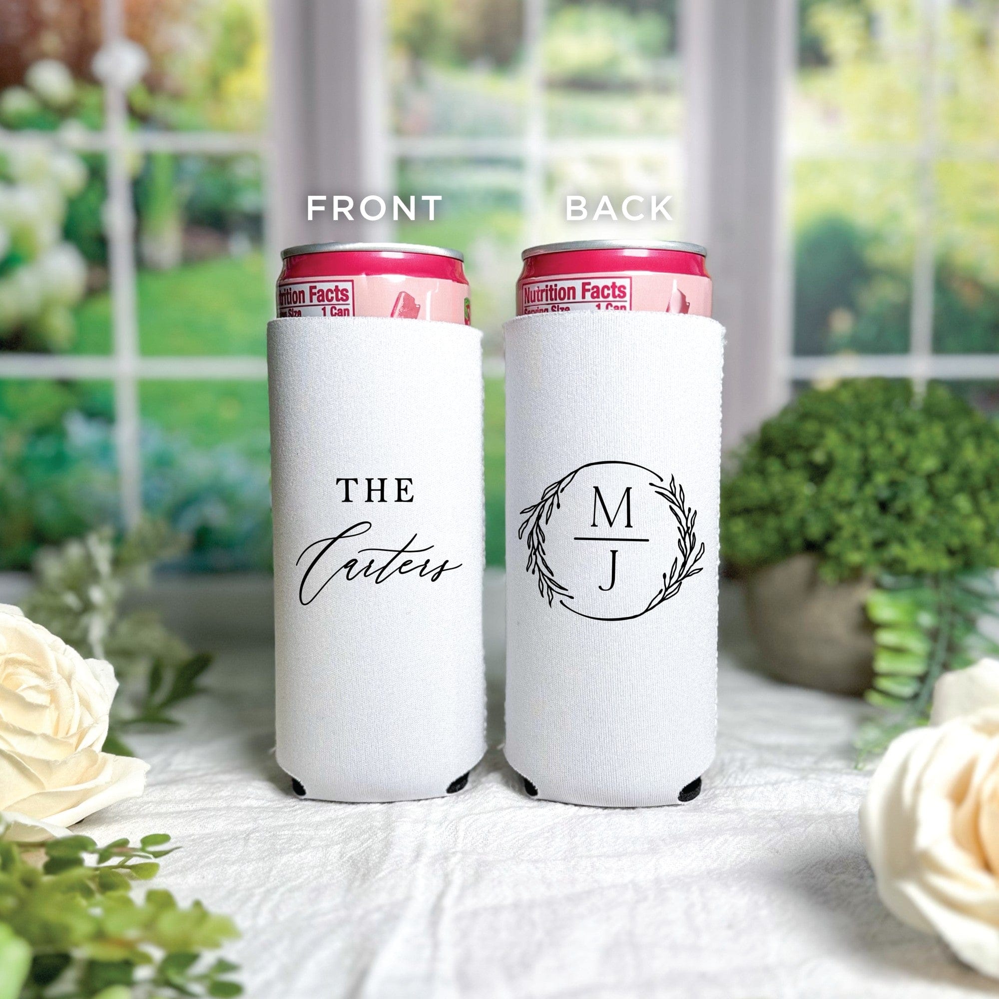 Two white neoprene slim can coolers for a wedding or event, one showcasing a custom family name graphic and the other a personalized initial monogram within a wreath design, holding pink beverage cans.