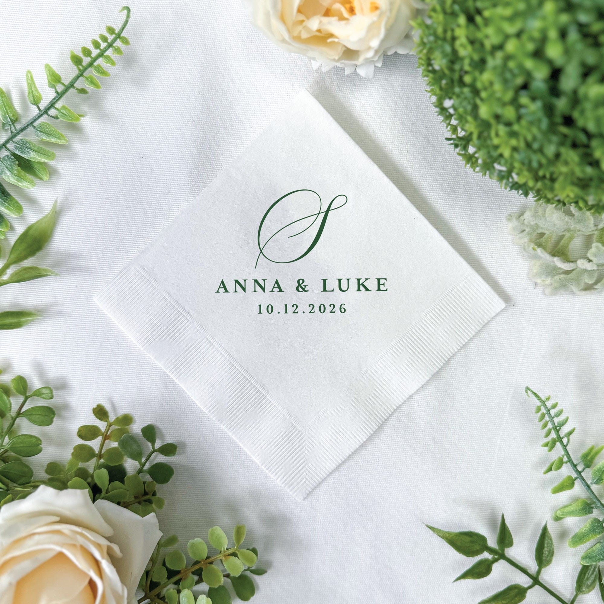 A white paper wedding napkin with an embossed border displays a custom dark green graphic element and multiple lines of personalized text, set on a white fabric background adorned with green foliage and light cream roses.