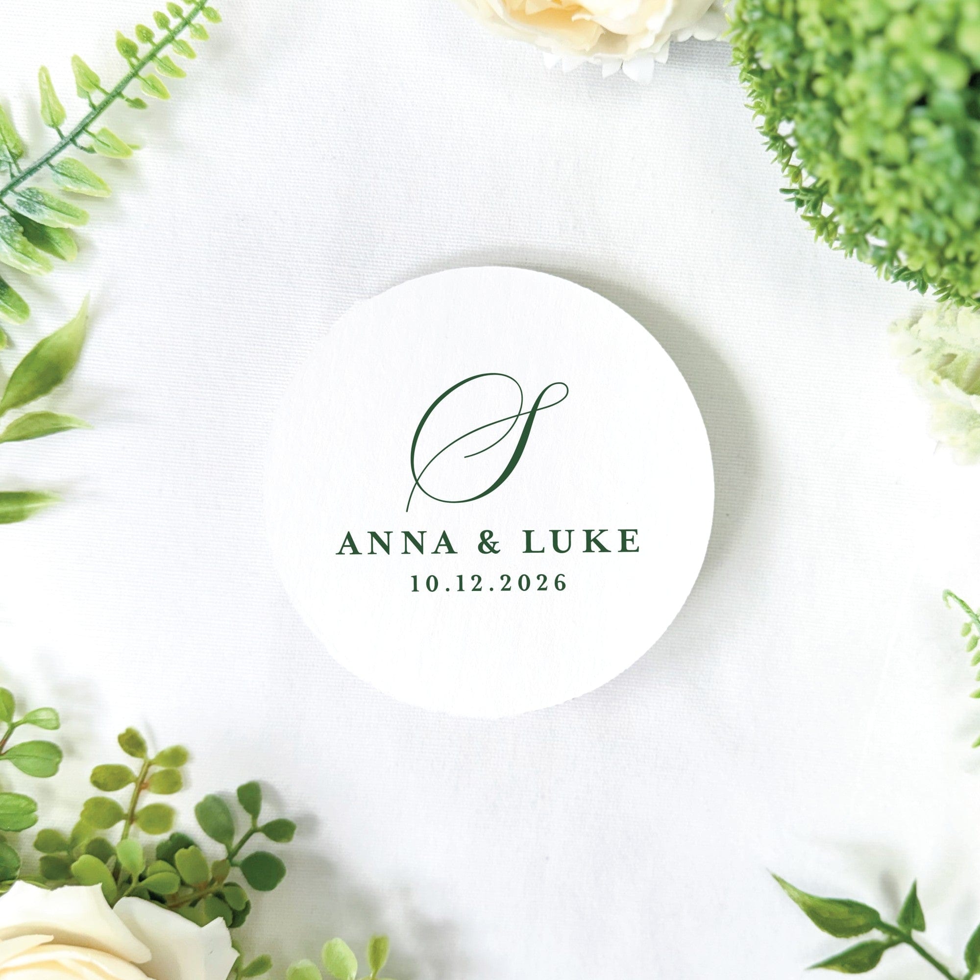 A white circular paper item featuring a personalized green script initial and custom text, designed for elegant wedding decor, presented on a white linen surface with green botanical elements and white flowers.