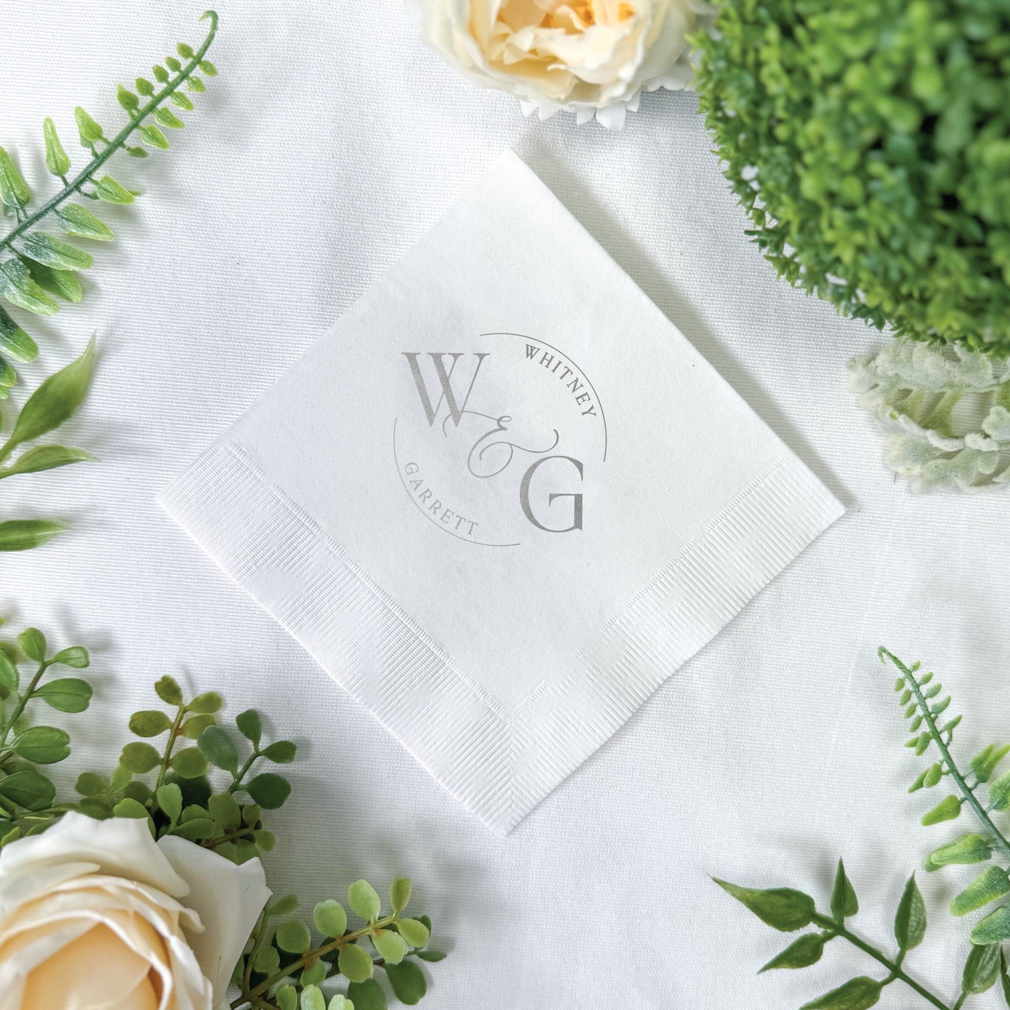 A folded white paper wedding napkin featuring a personalized circular gray monogram design, ideal for guest use or as elegant event decor.