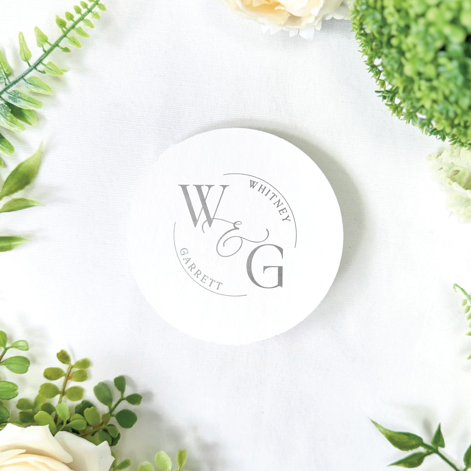 A white round paper coaster with a gray custom monogram design featuring initials and names, ideal for personalized wedding event decor.