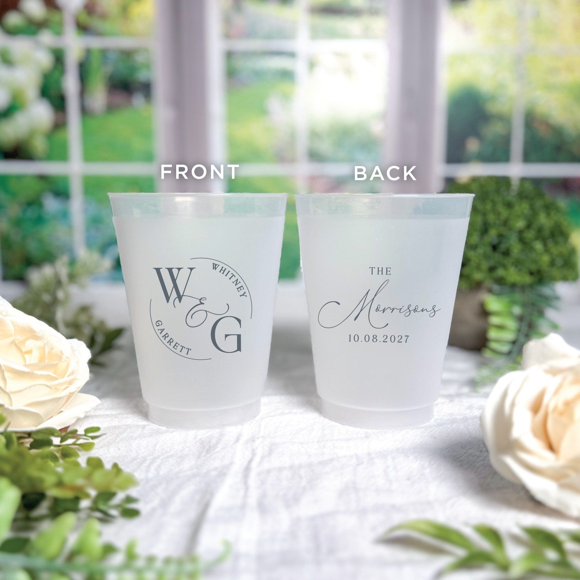 Two frosted white plastic wedding cups are displayed, one featuring a front view with a circular custom monogram design and the other showing a back view with a personalized calligraphic family name, both printed in dark gray and set on a white tablecloth with floral accents.