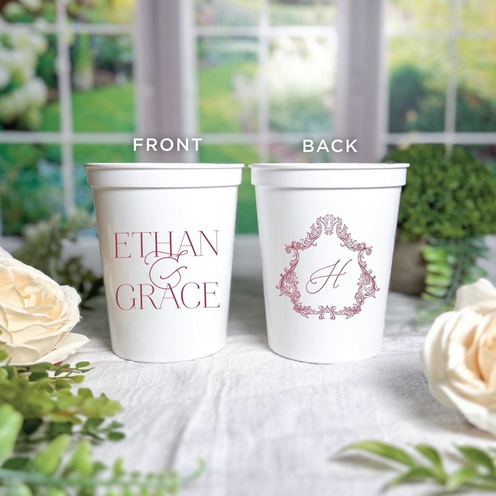 Two white plastic disposable cups, personalized for a wedding, feature a maroon print with two names joined by an ampersand on the front and an elegant framed initial on the back.