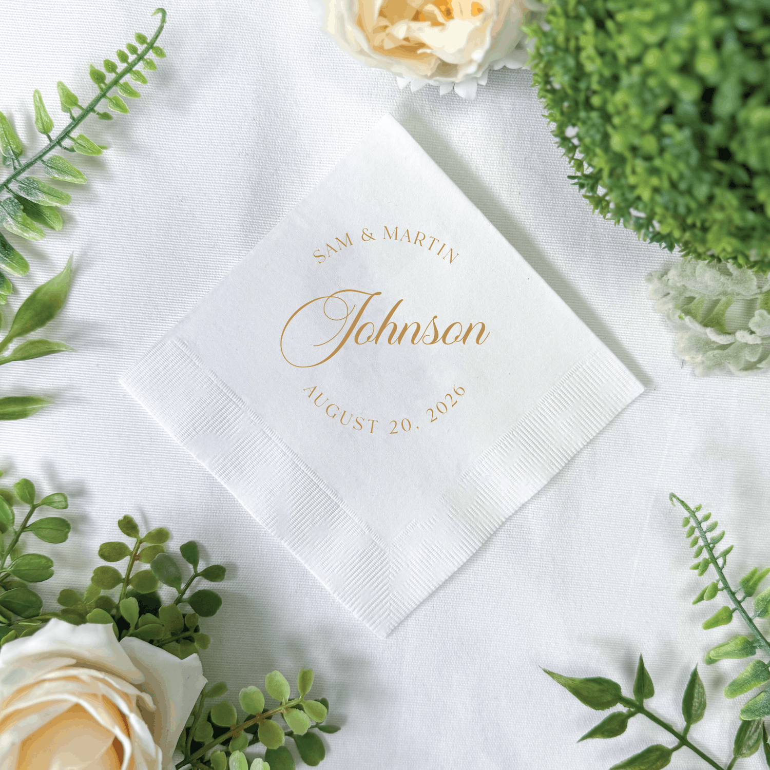 White cocktail napkin with gold foil text reading "Johnson" and wedding date, surrounded by greenery and cream roses.