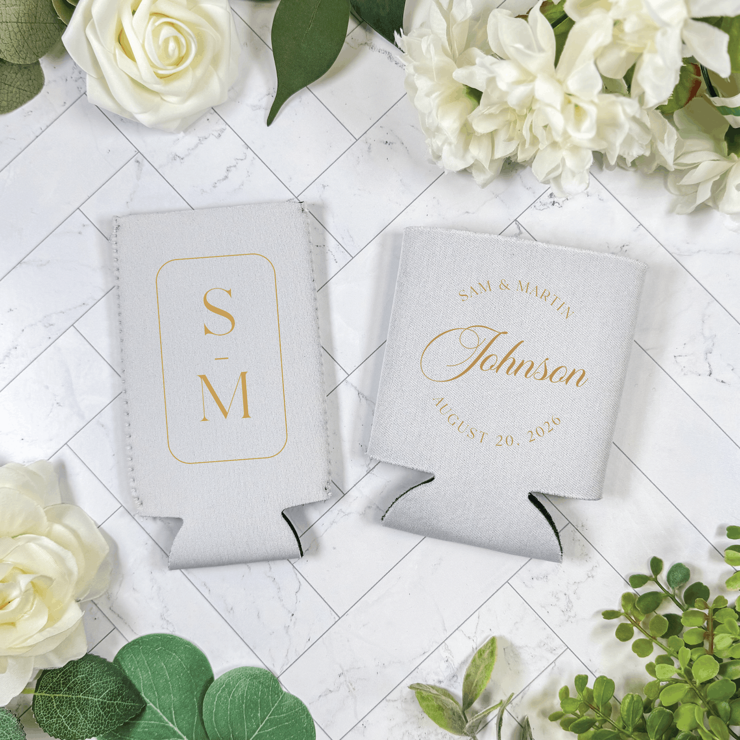 White can coolers with gold monogram and couple's name "Johnsen" wedding date, surrounded by white roses and greenery on marble background
