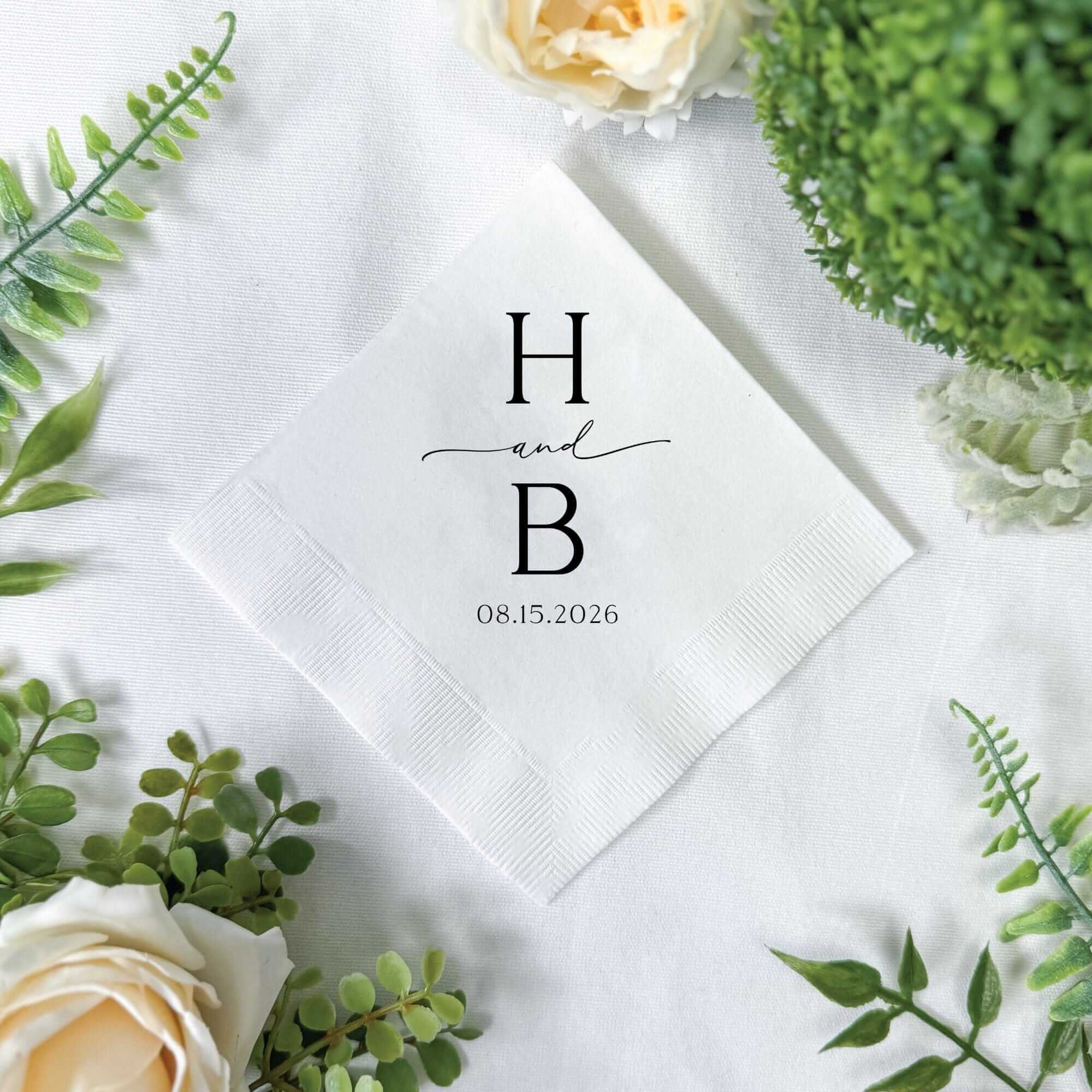 A crisp white paper wedding napkin, featuring elegant black personalized lettering, is beautifully displayed on a white linen-like surface, adorned with delicate green foliage and soft cream roses for sophisticated event decor.