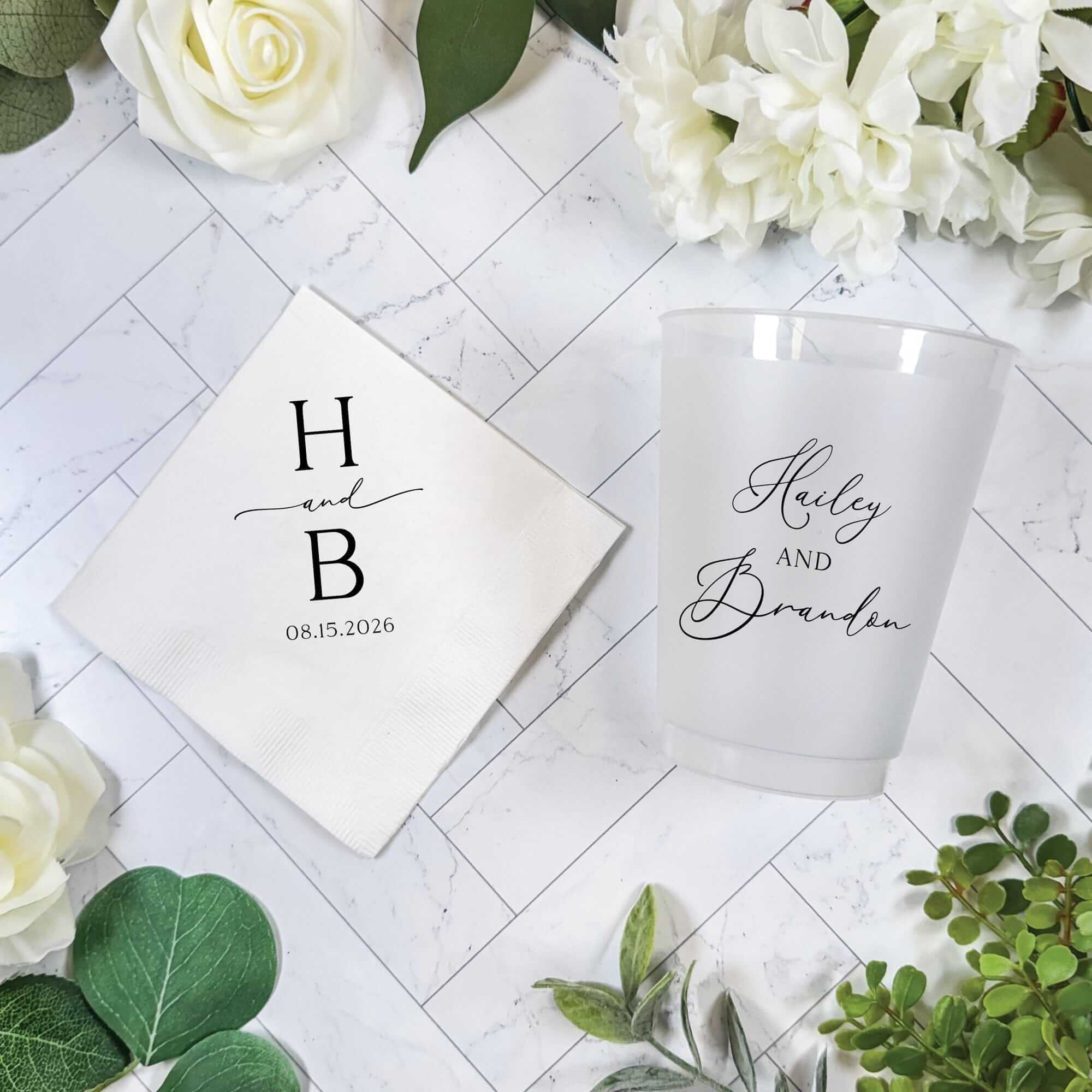 A white textured paper napkin and a frosted translucent plastic cup, both custom-printed for a wedding or special event, are artfully arranged on a white marble background with decorative white roses and green foliage.