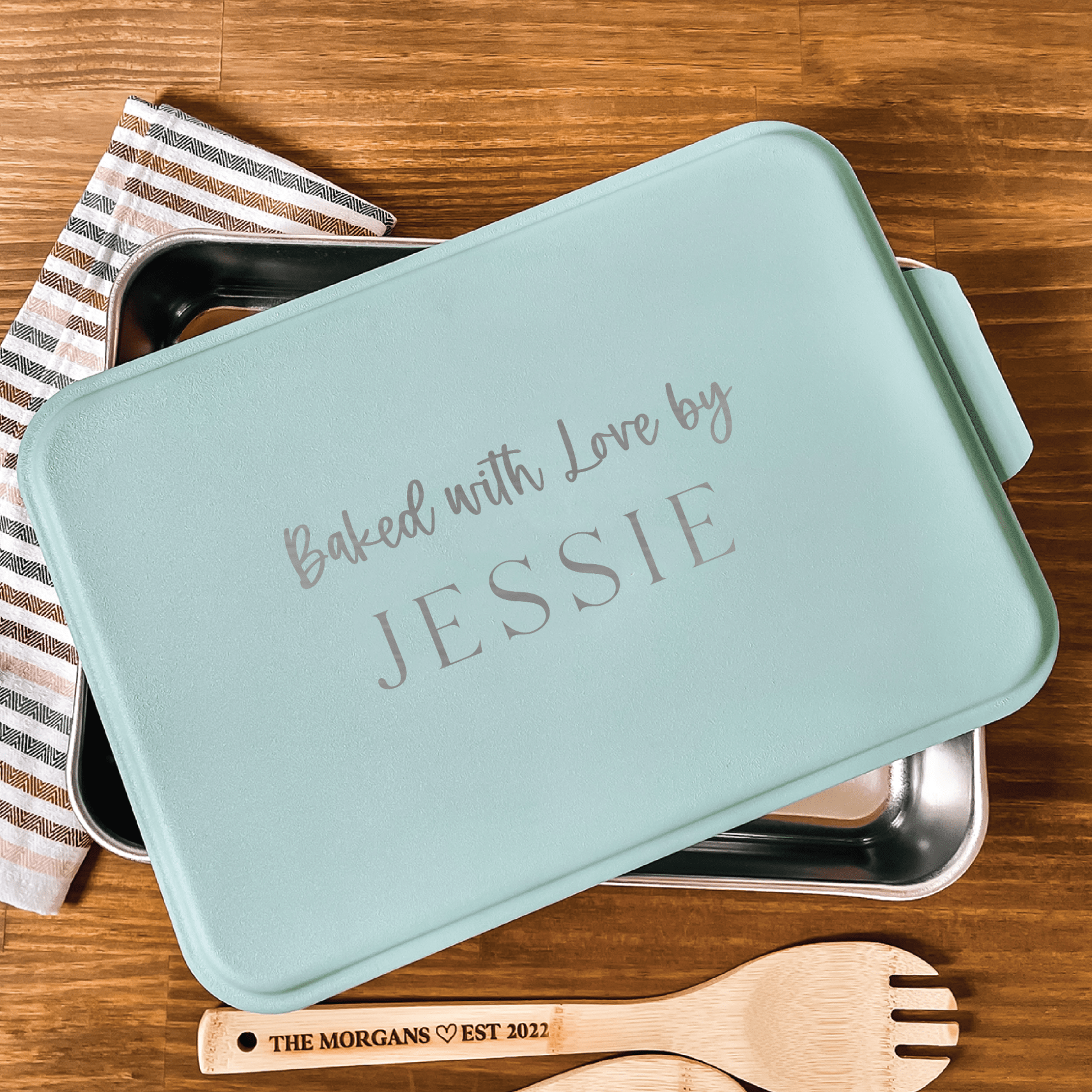 Light blue cake pan with "Baked with Love by JESSIE" engraved on lid, displayed with striped cloth, wooden serving utensils, and personalized wooden spoons.