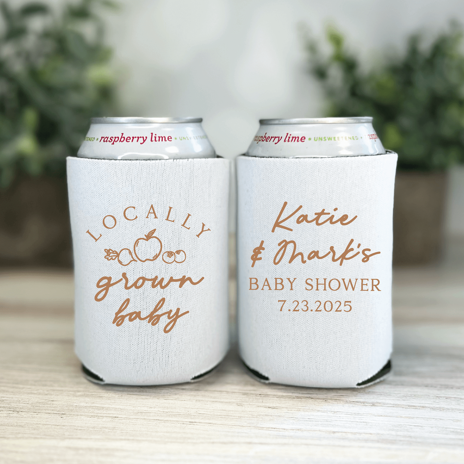 Two white can coolers with rose gold text, one reading "Locally Grown Baby" with fruit design, the other personalized "Katie & Mark's Baby Shower 7.23.2025"