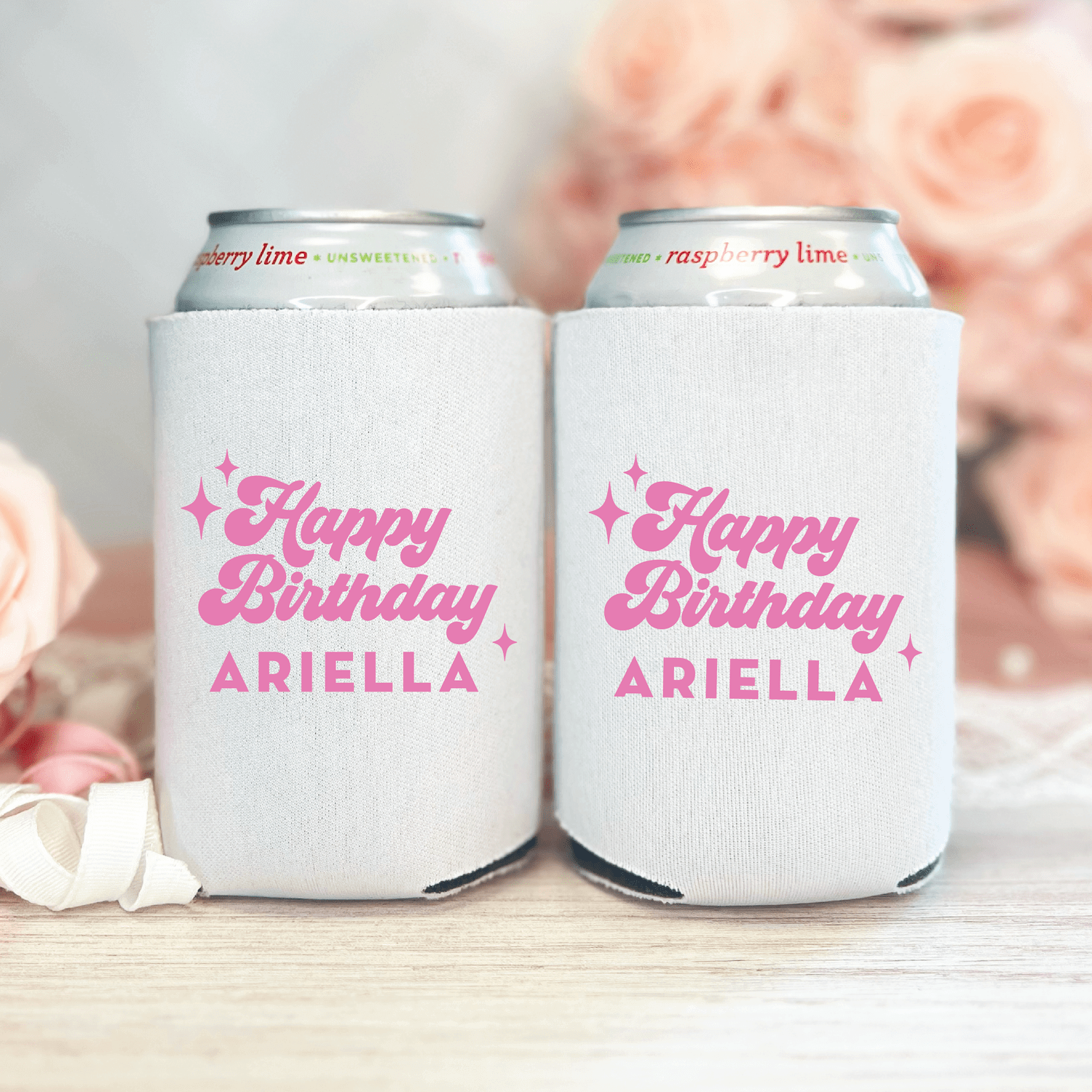 Two white can coolers with pink "Happy Birthday ARIELLA" text and star designs, holding beer cans with blurred flowers in background.