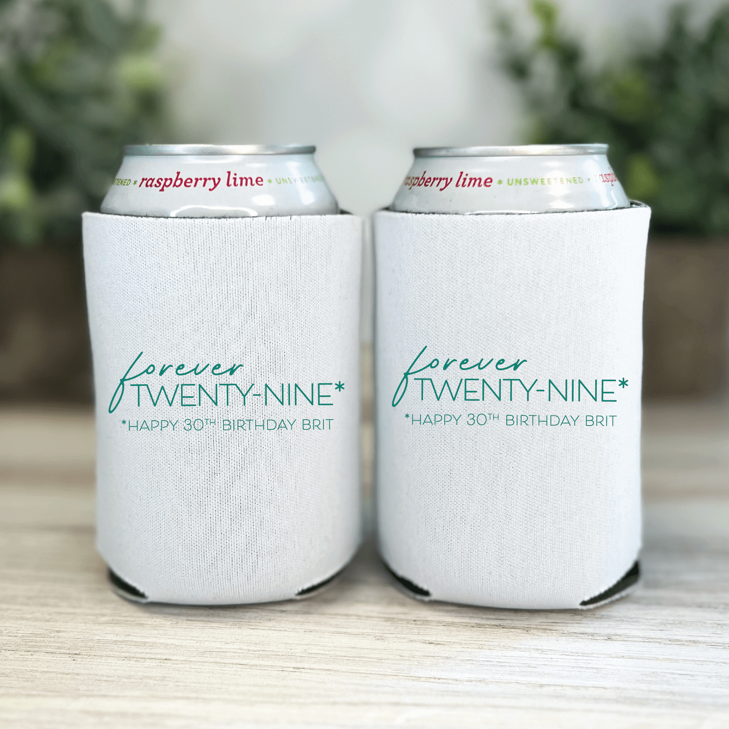 White can coolers with "forever twenty-nine" and "happy 30th birthday brit" personalized text in teal, holding canned beverages outdoors