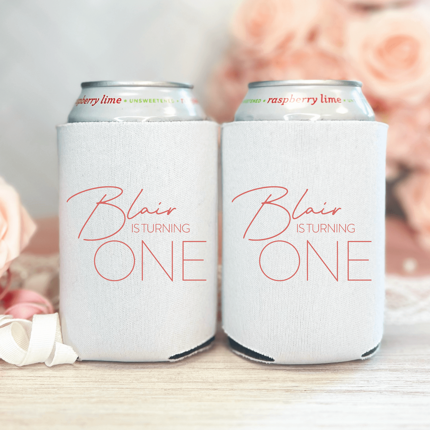 White can coolers with "Blair is turning ONE" in pink script text, holding cans with blurred flowers in background