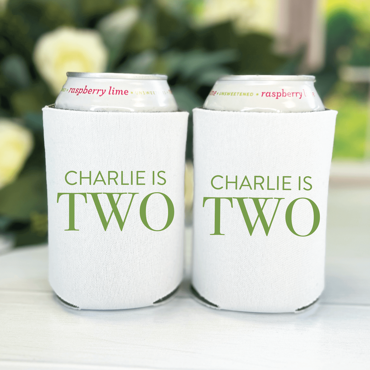 Pair of white can coolers with "CHARLIE IS TWO" in green text, holding cans with raspberry and lime flavor labels