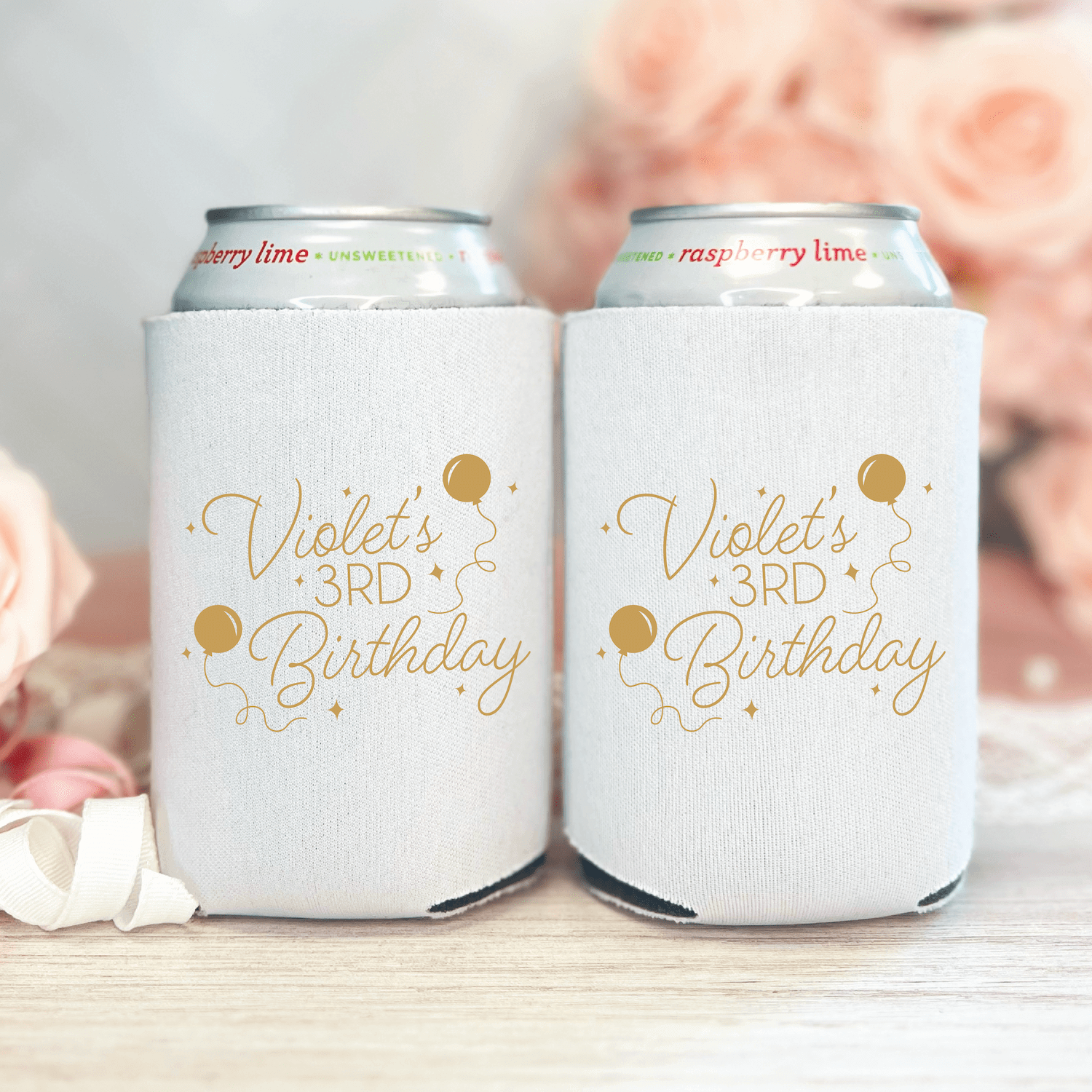 White can coolers with gold "Violet's 3rd Birthday" text and confetti design, displayed in pairs with blurred floral background.
