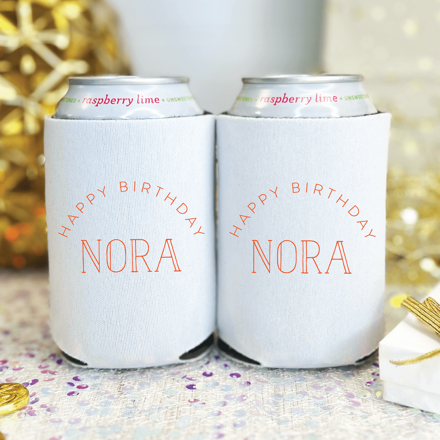 White can coolers with "Happy Birthday Nora" in coral text, displayed as a pair with festive gold and purple party decorations in background