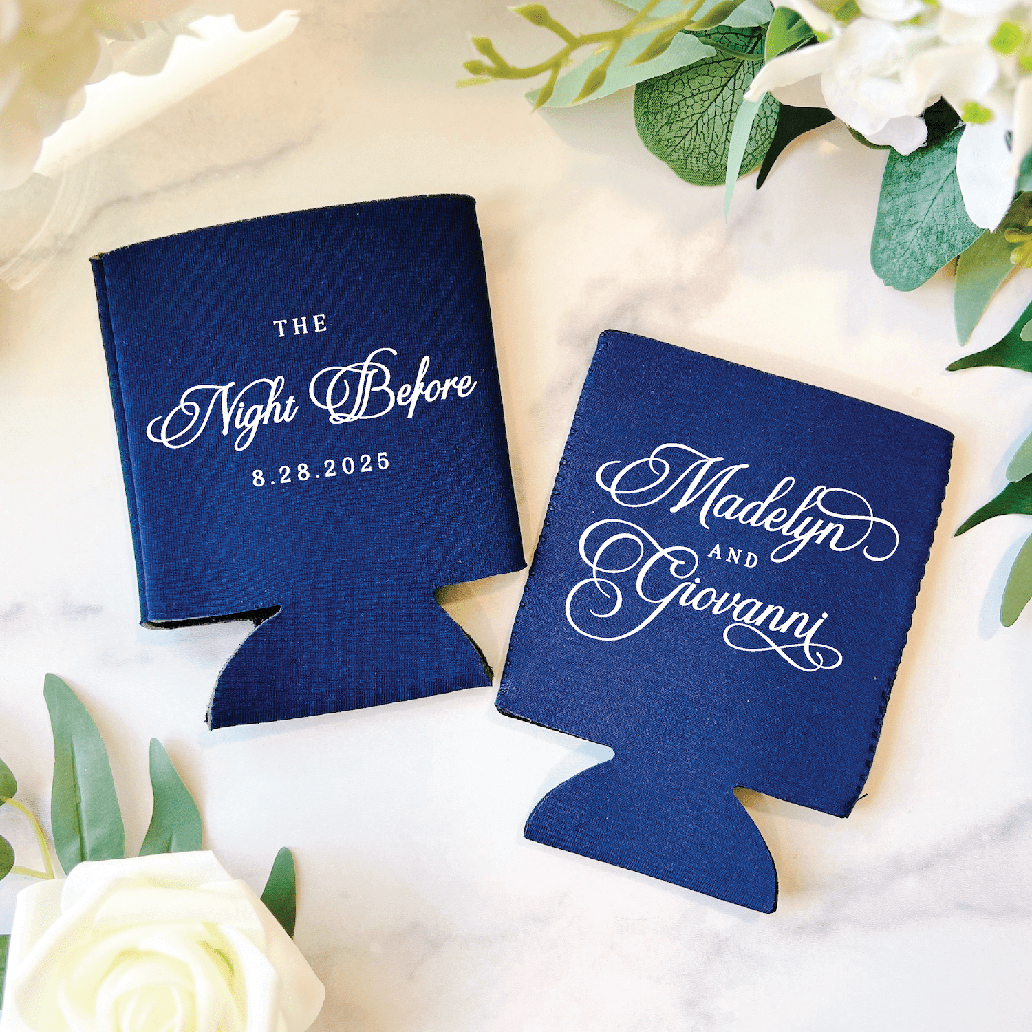 Navy can coolers with white script text reading "The Night Before" with date and couple's names, surrounded by white flowers and greenery