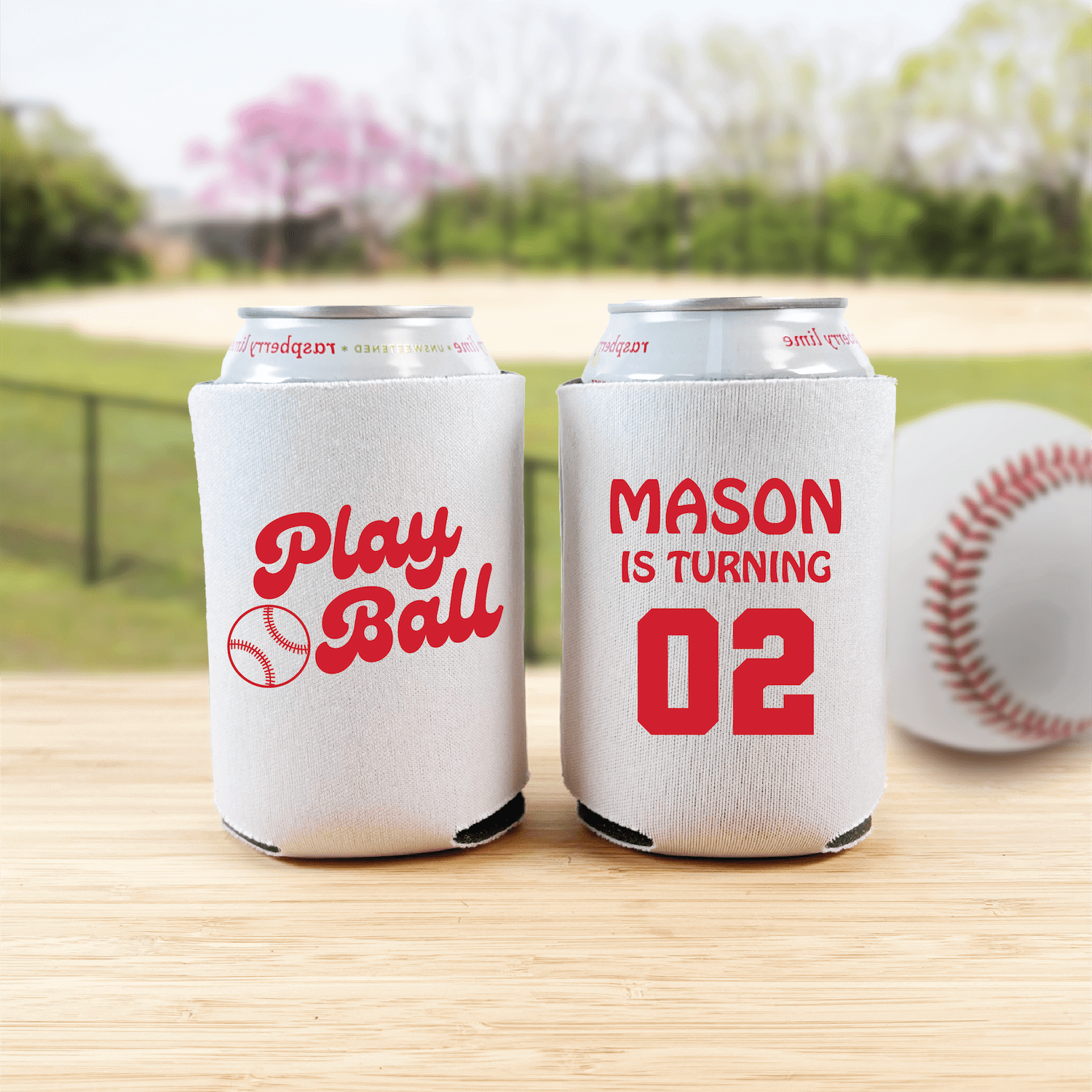 Two white can coolers with red baseball-themed designs on a wooden table, one reading "Play Ball" with baseball graphic, the other personalized "Mason is turning 02"