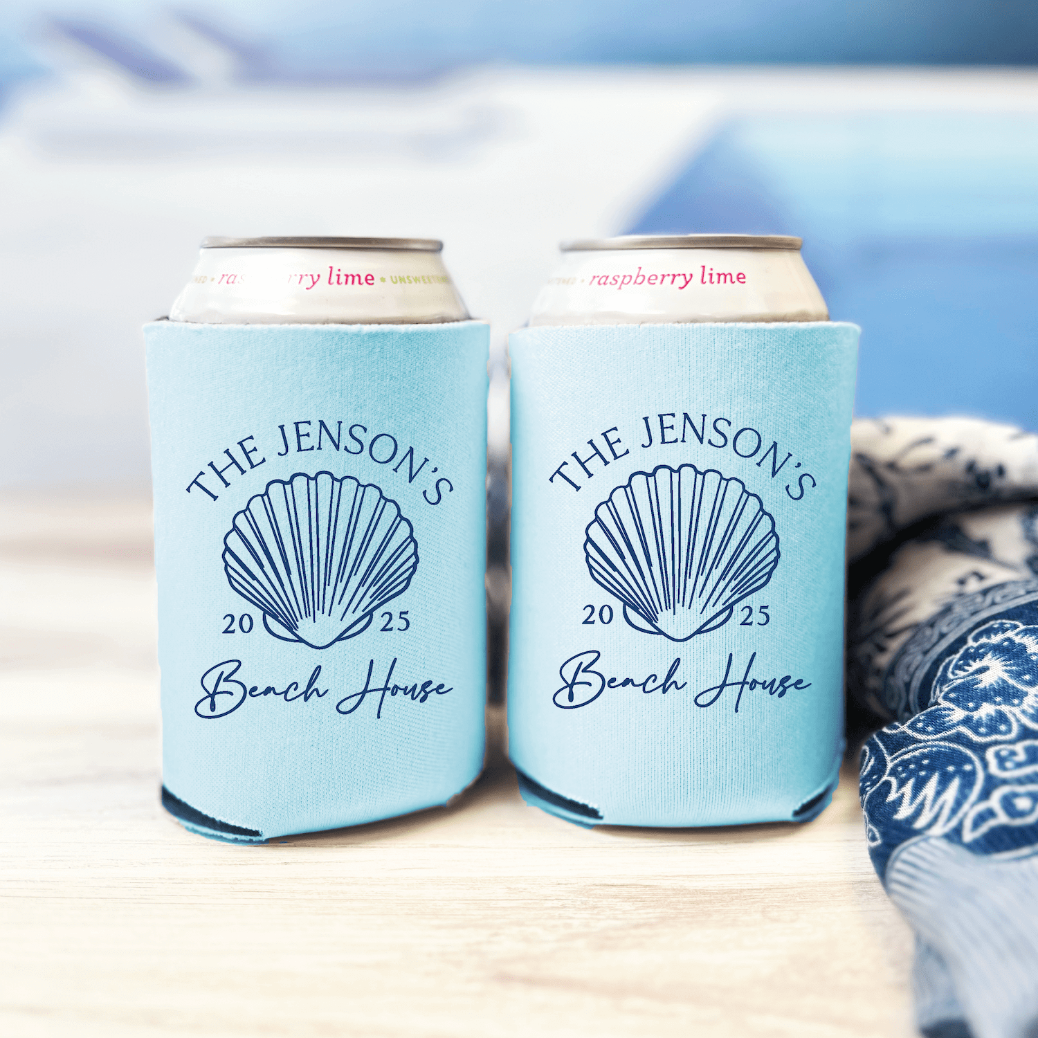 Light blue can coolers with seashell design and "The Jensons Beach House 2025" text in navy, displayed with personalized drink cans on a wooden surface.